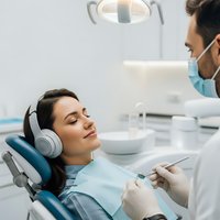 Young woman relaxing with headphones during dental treatment while dentist examines teeth in modern clinic for stress-free oral healthcare.