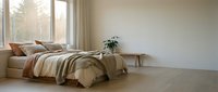 Cozy bedroom with unmade bed, warm neutral bedding, wooden nightstand with plant, large window with natural light and curtains.