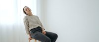 Young woman in casual beige sweater and dark pants sitting relaxed on wooden stool against white wall in minimalist interior space.