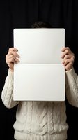 Man in beige cable knit sweater holding blank white paper sign against dark background for mockup and advertising purposes.