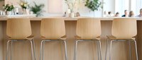 Young woman with light skin tone, wearing casual attire, sits at modern wooden bar counter with four empty chairs. Bright, minimalist kitchen interior with potted plants and natural light. Suitable for hospitality, interior design, and lifestyle content.