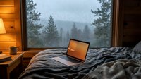 Cozy cabin bedroom with laptop on bed overlooking misty forest landscape through large window, warm wooden interior creating peaceful remote work atmosphere.