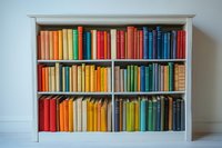 Colorful books arranged by spectrum colors in white wooden bookshelf against light wall, creating rainbow effect in home library or reading room setting.