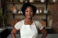 Young African American woman wearing white apron and beige shirt standing confidently in modern rustic kitchen with exposed brick wall and wooden shelving background.