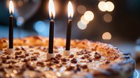 Birthday cake with lit candles against blurred bokeh lights background, sprinkled with chocolate crumbs and decorated with cream frosting.