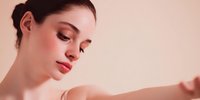 Young Caucasian woman with natural makeup and hair in bun looking down thoughtfully against soft beige background, showcasing delicate features and glowing skin.
