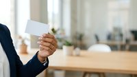 Businesswoman in formal navy blazer holding blank white business card in modern office environment with wooden desk and blurred background for corporate branding and professional networking.