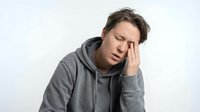 Young Caucasian man in gray hoodie experiencing severe headache, holding head in pain with eyes closed, stress and migraine concept for health awareness.