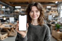 Young woman with curly hair showing blank smartphone screen in woodworking workshop, wearing gray sweater, carpentry studio background.