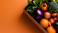 Fresh organic vegetables and fruits in wooden box on orange background, including red onion, tomatoes, carrots, oranges and broccoli. Healthy eating concept.