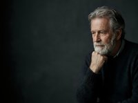 Senior man with gray hair and beard, wearing dark sweater, hand on chin in thoughtful pose against dark background. Suitable for business, healthcare, and lifestyle editorial use.