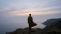 Man in casual clothing standing on coastal cliff at sunset, gazing toward ocean horizon. Suitable for travel, lifestyle, and inspirational content.