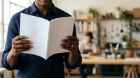 Man with dark skin tone wearing navy blue shirt reading open magazine or brochure in bright office workspace with plants and shelves, suitable for business, education, and publishing content.
