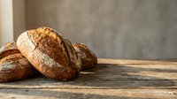 Freshly baked artisan bread loaves with golden crust on rustic wooden table against textured wall background for bakery and food photography.