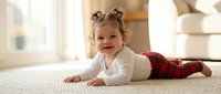 Happy toddler girl with pigtails lying on carpet in cozy home interior wearing white top and red plaid pants smiling at camera for family lifestyle content.