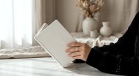 Woman in black sweater reading open book in cozy bedroom with dried flowers and soft natural lighting creating peaceful morning atmosphere.