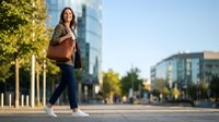 Young professional woman walking confidently through modern urban business district during golden hour sunset with leather handbag and casual attire.