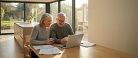 Senior couple using laptop together at home reviewing documents and planning finances in modern kitchen with large windows.