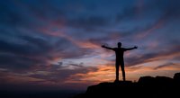 Male silhouette standing on mountain peak with arms raised celebrating success against dramatic sunset sky with purple and orange clouds for motivation.