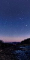 Starry night sky over rocky terrain with pink horizon glow and scattered stars reflecting in wet ground surface, creating dreamy atmospheric landscape.