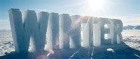 Large ice letters spelling WINTER stand on a snowy landscape under bright sunlight with lens flare, evoking cold season themes.