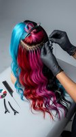 Hairstylist wearing black gloves applies vibrant blue, magenta, and red hair extensions to long wavy hair, demonstrating professional color blending and styling techniques for salon services.