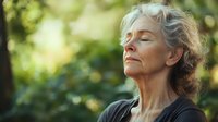 Senior woman with gray hair meditating outdoors in nature with eyes closed and peaceful expression, sunlit bokeh background creates serene atmosphere.
