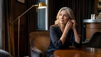 Thoughtful middle-aged Caucasian businesswoman in dark blazer sitting at wooden table in elegant office interior with warm lighting and modern decor.