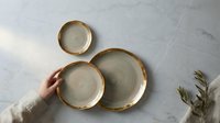 Ceramic bowls with gold rim held by hand on marble surface with dried botanicals, suitable for tableware, home decor, and lifestyle photography.