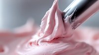 Creamy pink frosting being piped from metal pastry tip, macro shot showing smooth swirled texture and soft pastel color against blurred background.