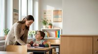 Young Asian mother helping elementary school age son with homework at wooden desk in bright modern home interior with natural lighting from windows.