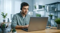Young man with dark hair and light skin, wearing headphones and casual shirt, working on laptop at wooden desk in modern office environment with plants and shelving. Suitable for customer service, tech support, remote work, and business communication concepts.