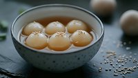 Traditional Asian sweet soup with glutinous rice balls in ceramic bowl, comfort food dessert with sesame seeds scattered around dark wooden surface.