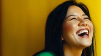 Young Asian woman with bright genuine smile laughing against vibrant yellow background, showing joy and happiness in casual green attire. Natural lifestyle portrait.