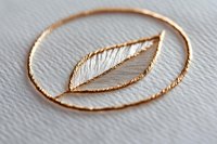 Delicate rose gold leaf pendant with white thread details on textured paper background, showcasing intricate craftsmanship in modern jewelry design.