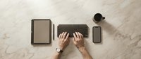 Professional workspace with hands typing on wireless keyboard surrounded by tablet, smartphone and coffee cup on marble surface for productivity and business.
