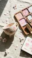 Watercolor painting supplies with floral teacup and pink paint palette on textured surface with scattered cherry blossom petals in warm natural sunlight.