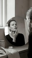 Middle-aged woman of Caucasian ethnicity examining her reflection in a round mirror, touching her face thoughtfully. Monochrome portrait for skincare, beauty, wellness, and self-care concepts.