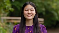 Young Asian woman with long dark hair smiling warmly outdoors in purple top against natural green background for lifestyle and portrait photography.