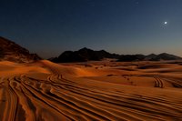 Desert sand dunes with vehicle tracks under starry night sky, dramatic mountain silhouettes against dark blue horizon, moonlit orange sandy landscape creates rippling patterns.