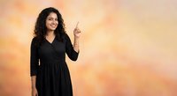 Smiling young Middle Eastern woman with curly hair in black dress pointing upward against warm orange gradient background for business presentations.