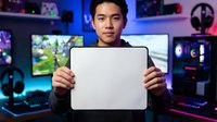 Young male gamer of East Asian descent holding blank laptop screen in neon-lit gaming room setup with multiple monitors and RGB lighting for tech promotion.