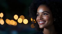 Young African American woman with natural curly hair smiling genuinely against blurred golden bokeh lights background, radiating joy and confidence in evening setting.