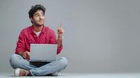 Young South Asian man in pink shirt sitting cross-legged with laptop, pointing upward with eureka moment expression against neutral gray background.