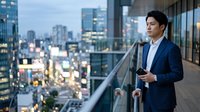 Young Asian male professional in navy blazer and light blue shirt standing on high-rise balcony overlooking sprawling cityscape, holding smartphone, urban business lifestyle.