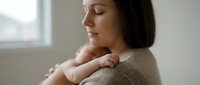 Tender moment as a young Caucasian mother holds her sleeping newborn baby against her shoulder, eyes closed in peaceful bonding.