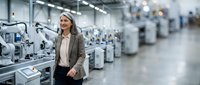 Middle-aged woman with blonde hair wearing business casual attire standing in modern manufacturing facility with industrial equipment and machinery in background.
