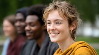 Young woman in yellow hoodie smiling with genuine joy while diverse group of friends gather outdoors on sunny day, expressing happiness and authentic friendship.