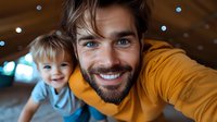 Young bearded Caucasian father in yellow sweater taking selfie with cheerful toddler son, both smiling at camera. Bokeh lights background creates warm atmosphere.