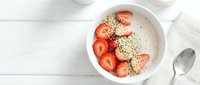 Overhead view of a white bowl containing creamy yogurt topped with fresh strawberry slices and granola, styled on white wooden surface with spoon for breakfast or healthy eating content.